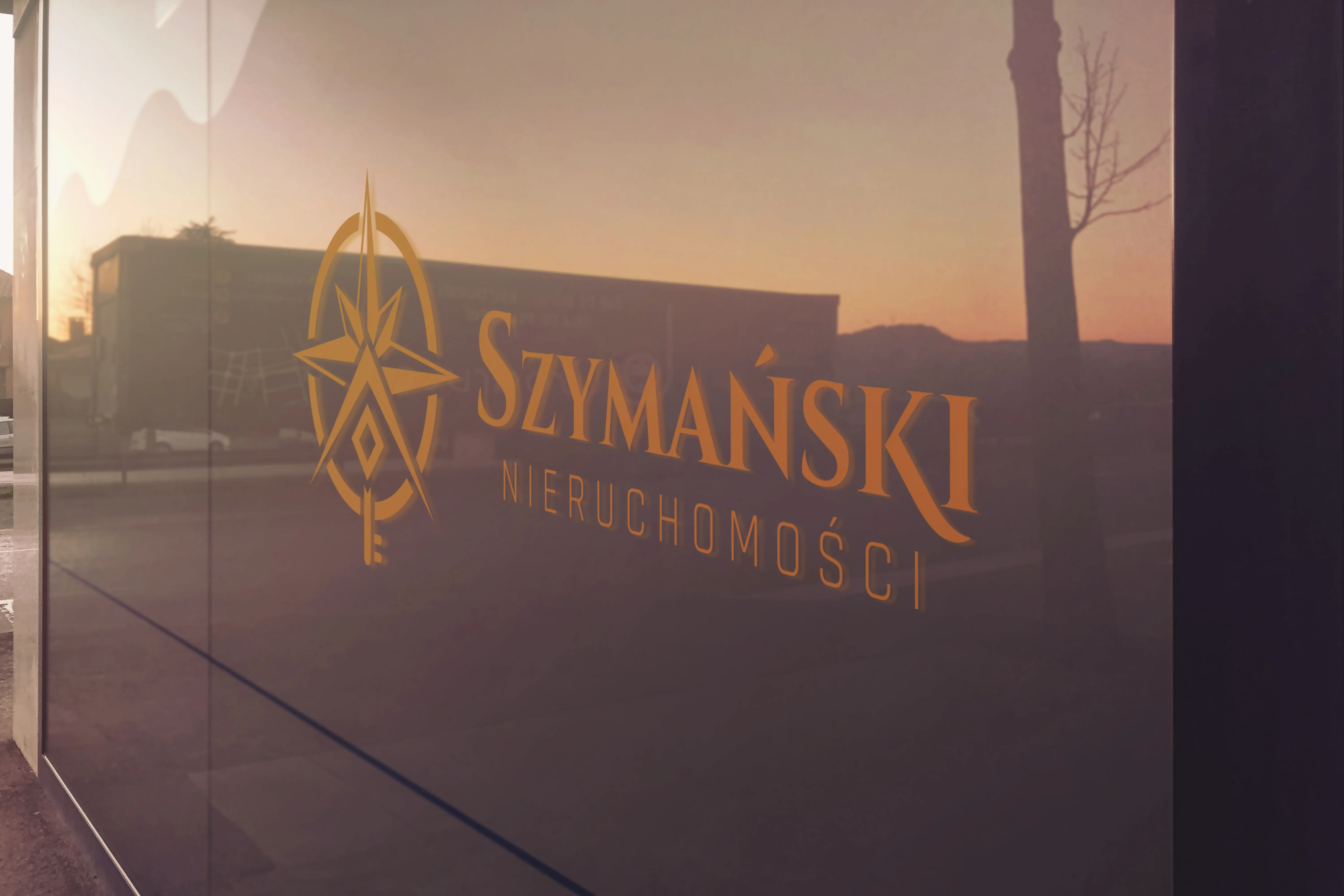 Wizualizacja logo Szymański Nieruchomości na szklanej witrynie biura o zachodzie słońca – złoty sygnet z różą wiatrów i logotyp